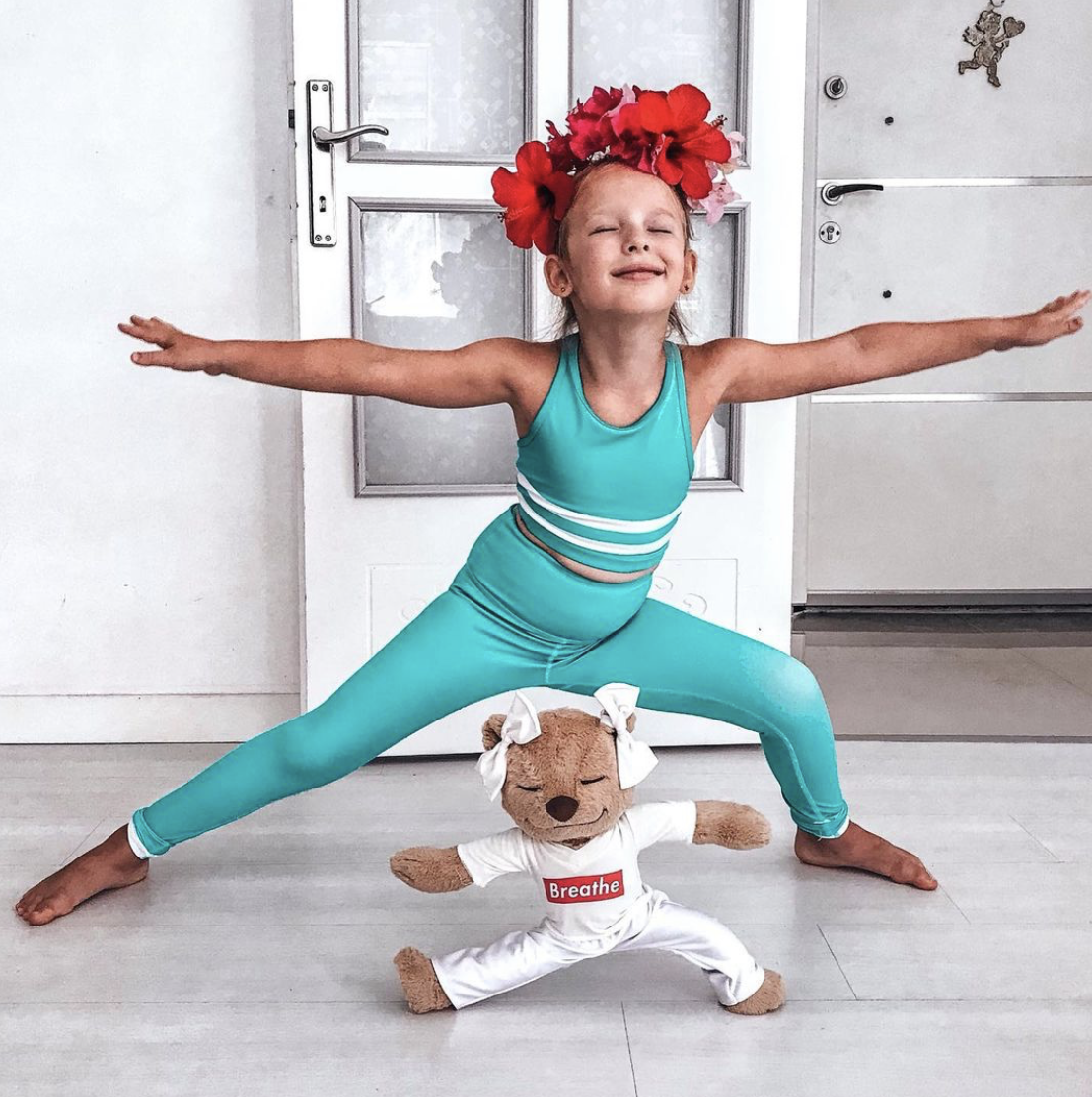 A young girl performing Warrior II (Virabhadrasana II) yoga pose with Meddy Teddy wearing white yoga pants and Breathe white T-shirt