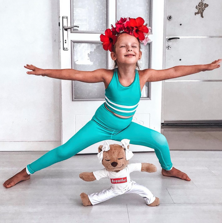 A young girl performing Warrior II (Virabhadrasana II) yoga pose with Meddy Teddy wearing white yoga pants and Breathe white T-shirt